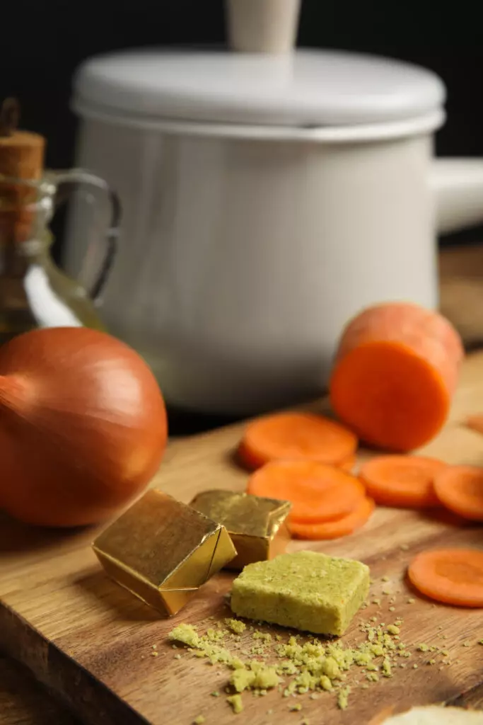 Close up of chicken bouillon cubes alongside carrots and onions on a cutting board