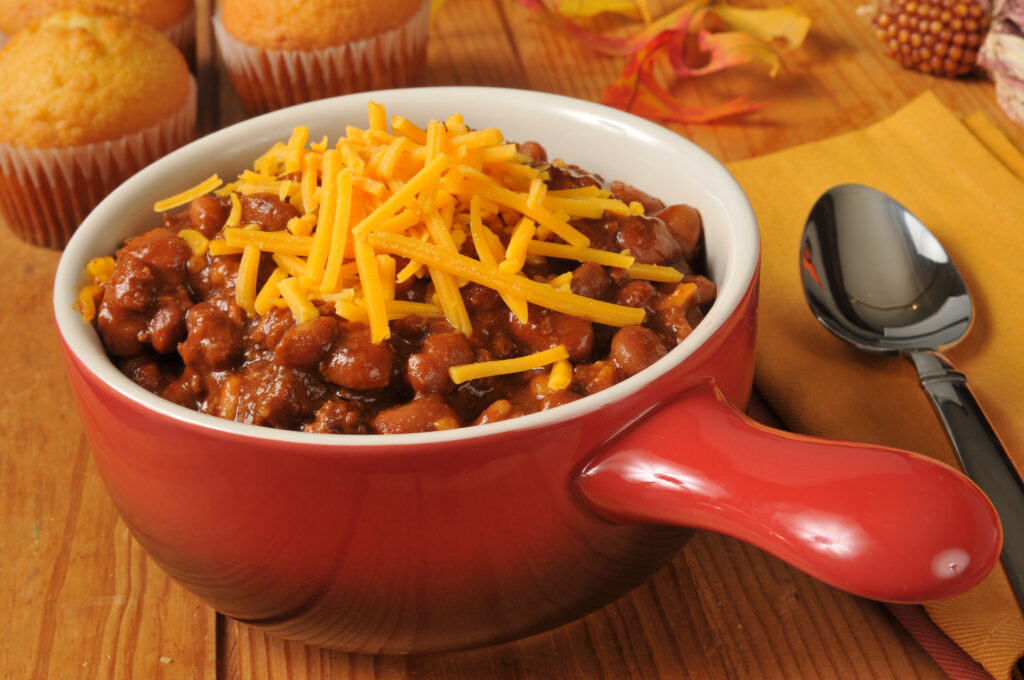 Close up shot of chili topped with cheese with corn bread muffins in the background.