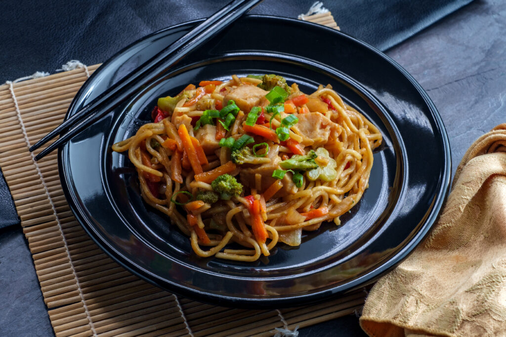 Chicken chow mein on a black plate on a bamboo placemat