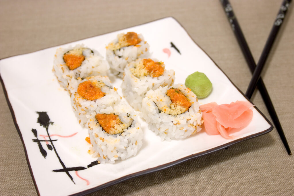 Spicy tuna roll arranged on a white plate with ginger and wasabi sauce with black chopsticks in the background. 