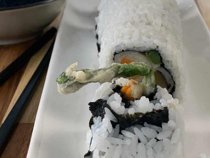 Vegetable tempura roll on a white sushi plate with a ramekin of soy sauce and chopsticks in the background.