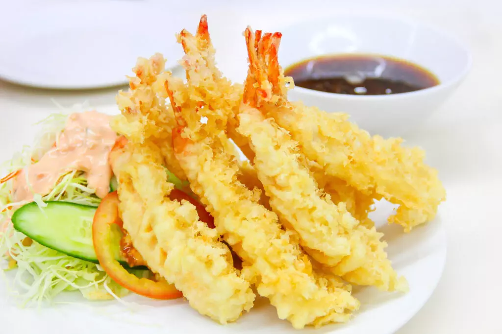 Close up of shrimp tempura with a tempura dipping sauce in the background and a salad.