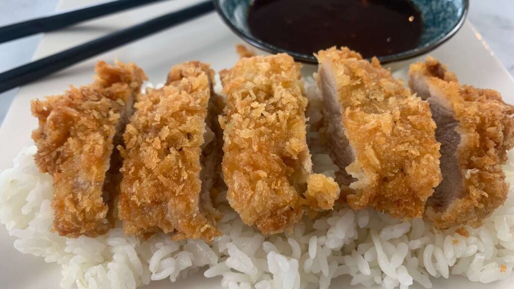 Chicken katsu on a bed of rice with katsu sauce and chopsticks in the background.