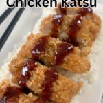 Close up of chicken katsu on a bed of rice with katsu sauce drizzled over the top. Served on a white plate with chopsticks and a ramekin of katsu sauce.