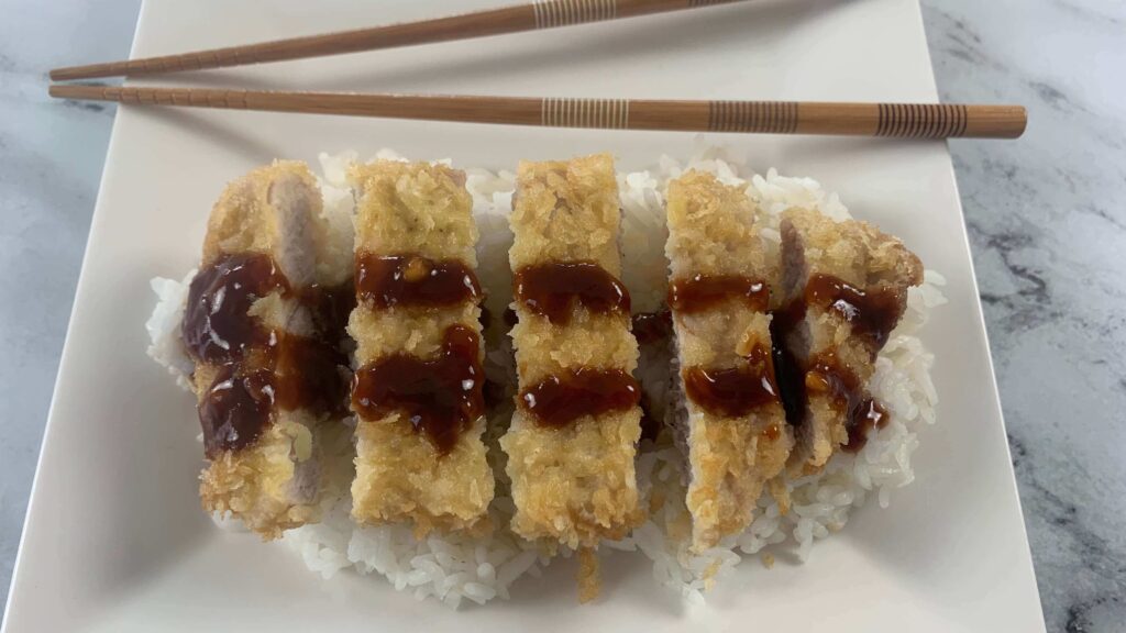 Tonkatsu pork sliced on a bed of rice covered with katsu sauce on a white plate.  Brown chopsticks in the background.