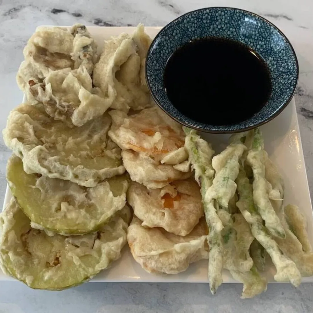 Vegetable tempura on a white plate with tempura dipping sauce in the background.