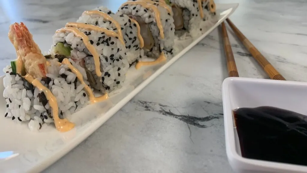 Dynamite rolls on a white sushi plate with a ramekin of unagi sauce and chopsticks in the background. 