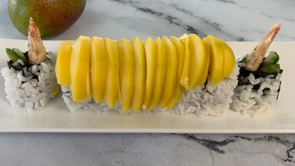 Mango sushi roll on a white sushi plate with a mango in the background.