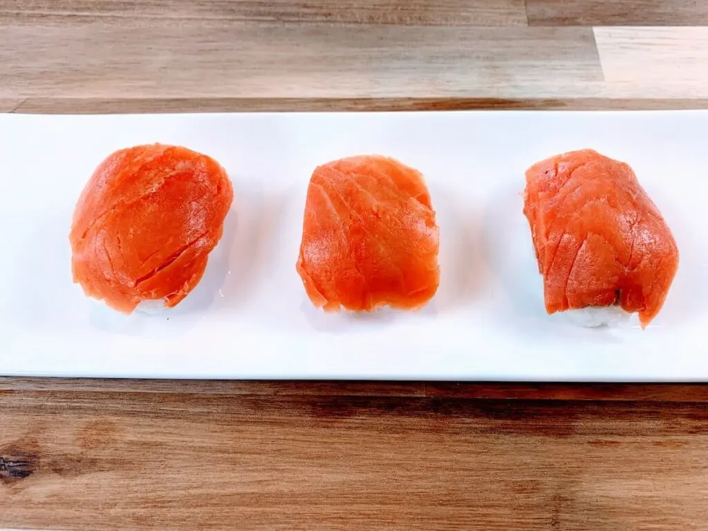 Salmon nigiri made with smoked salmon on a white sushi plate placed on a wood cutting board.
