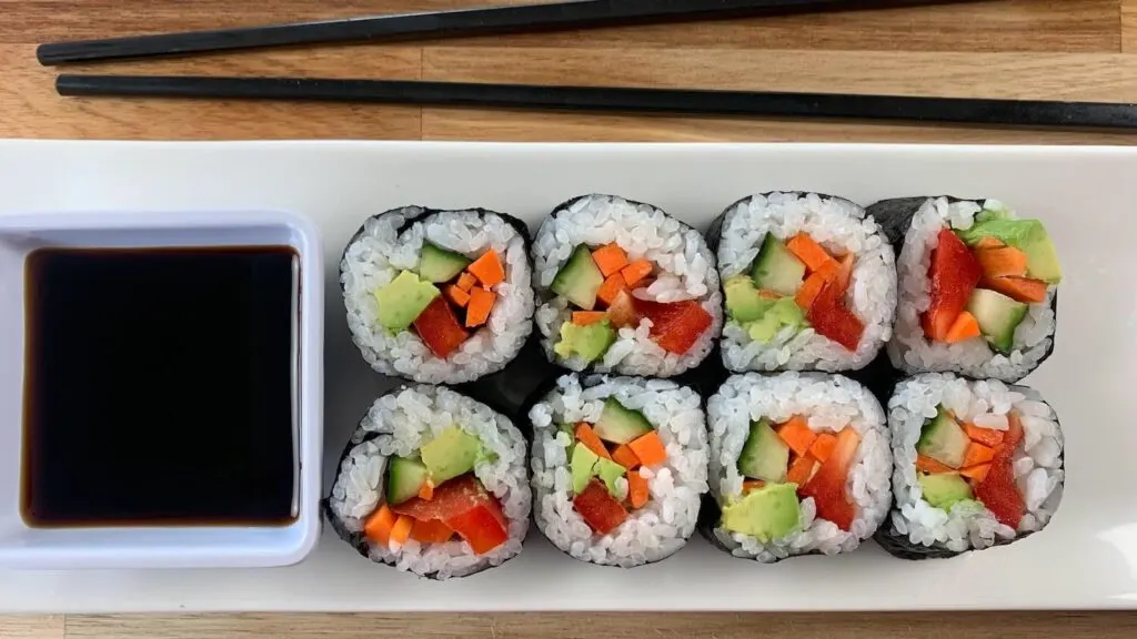 Vegetable roll sushi arranged on a white sushi plate with a ramekin of soy sauce and black chopsticks in the background. 