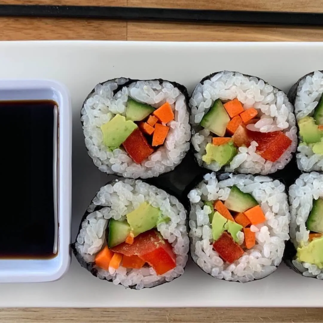 Close up of vegetable roll sushi on a white plate with a ramekin of soy sauce and a chopstick in the background.