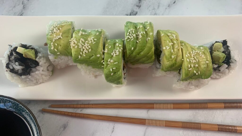 Caterpillar roll garnished with sesame seeds on a white sushi platter with chopsticks and soy sauce in the background. 