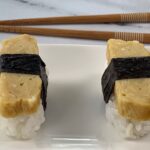 Tamago sushi square pic on a white plate with brown chopsticks in the background.