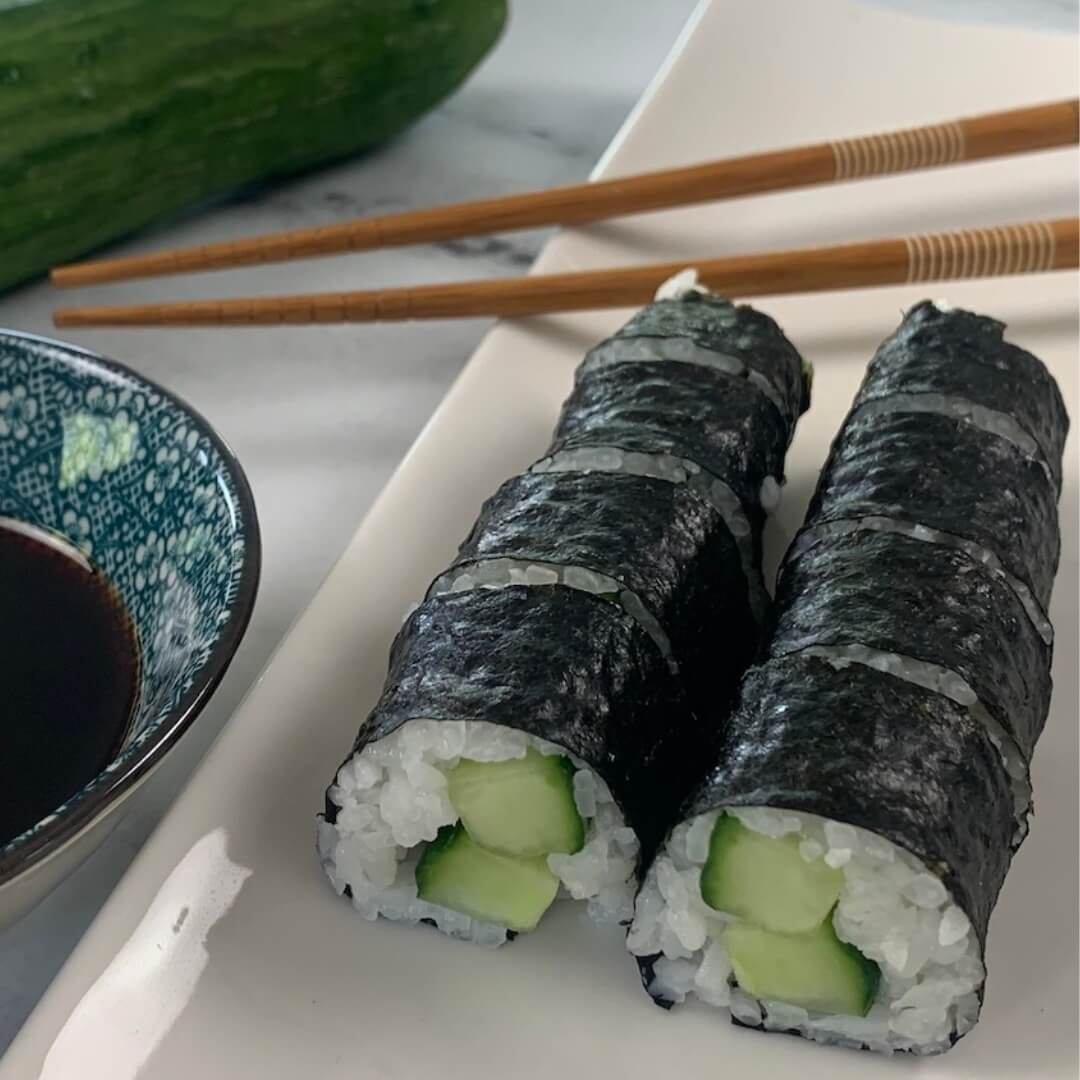 Cucumber rolls on a white plate with a ramekin of soy sauce, chopsticks, and a cucumber in the background.