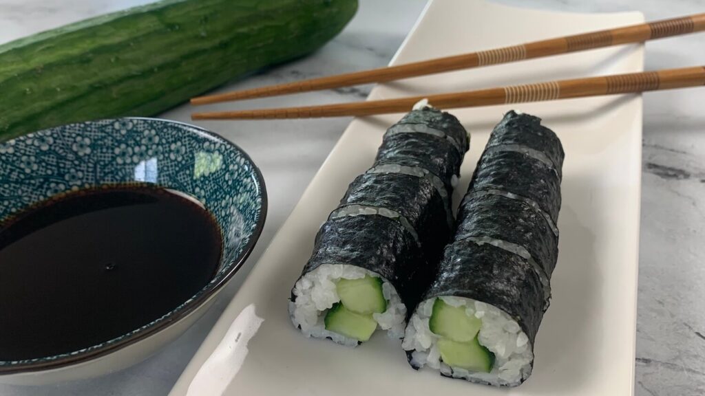 Cucumber roll (kappa maki) on a white sushi platter with chopsticks, a cucumber, and a ramekin of soy sauce in the background.