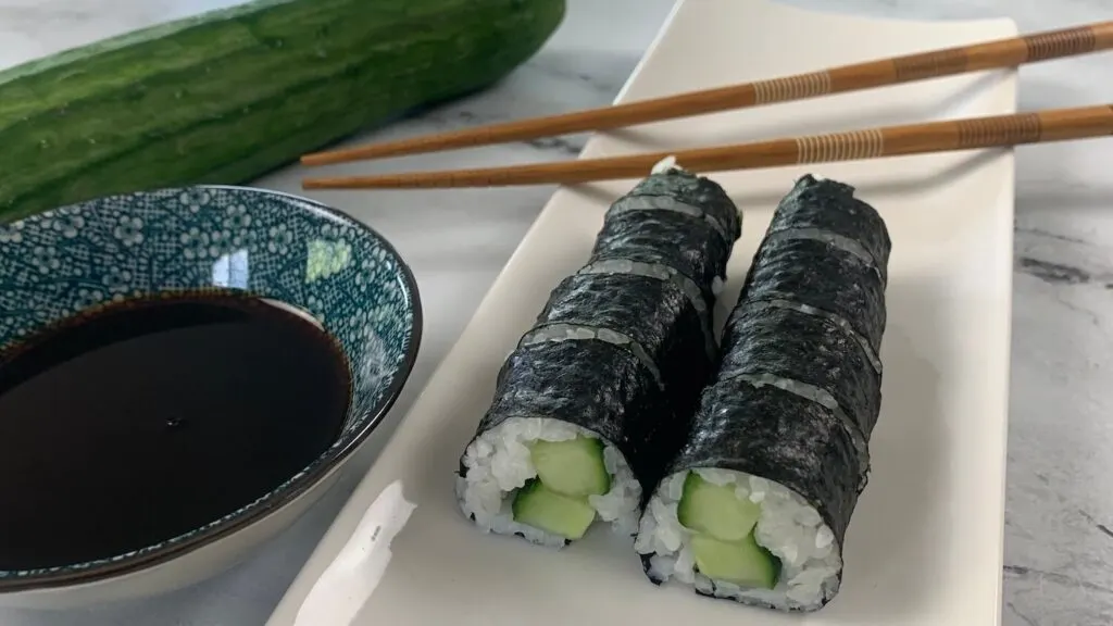 Cucumber roll (kappa maki) on a white sushi platter with chopsticks, a cucumber, and a ramekin of soy sauce in the background.