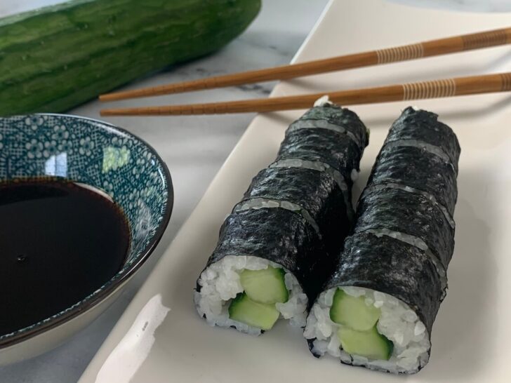 Cucumber roll (kappa maki) on a white sushi platter with chopsticks, a cucumber, and a ramekin of soy sauce in the background.