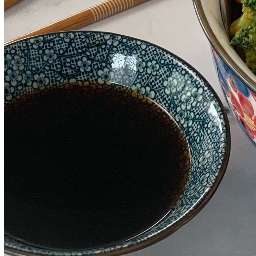 Teriyaki Sauce in a Japanese ramekin with a bowl of chicken teriyaki and chopsticks in the background.