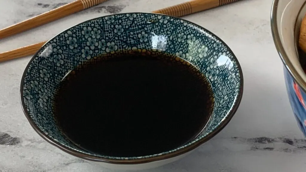 Horizontal pic of teriyaki sauce in a Japanese ramekin with chopsticks and a chicken teriyaki bowl in the background.