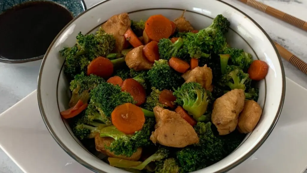 Horizontal pic of a chicken teriyaki bowl with teriyaki chicken, carrots, broccoli and rice.  Served in a Japanese bowl on a white plate with chopsticks and a ramekin of teriyaki sauce in the background.