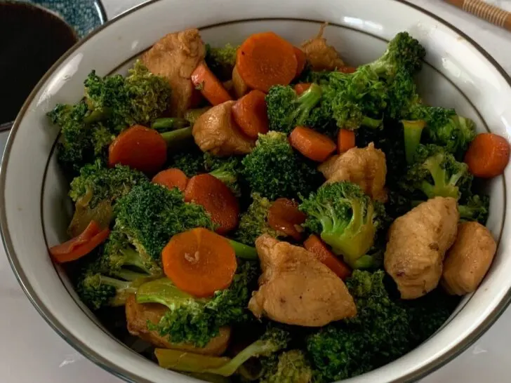 Horizontal pic of a chicken teriyaki bowl with teriyaki chicken, carrots, broccoli and rice. Served in a Japanese bowl on a white plate with chopsticks and a ramekin of teriyaki sauce in the background.