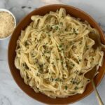 Alfredo sauce over fettuccini square pic garnished with diced parsley with a ramikin of parmesan cheese in the background.