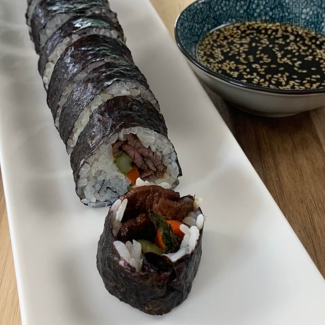 Beef roll sushi on a white sushi platter with a ramekin of yakiniku sauce in the background.