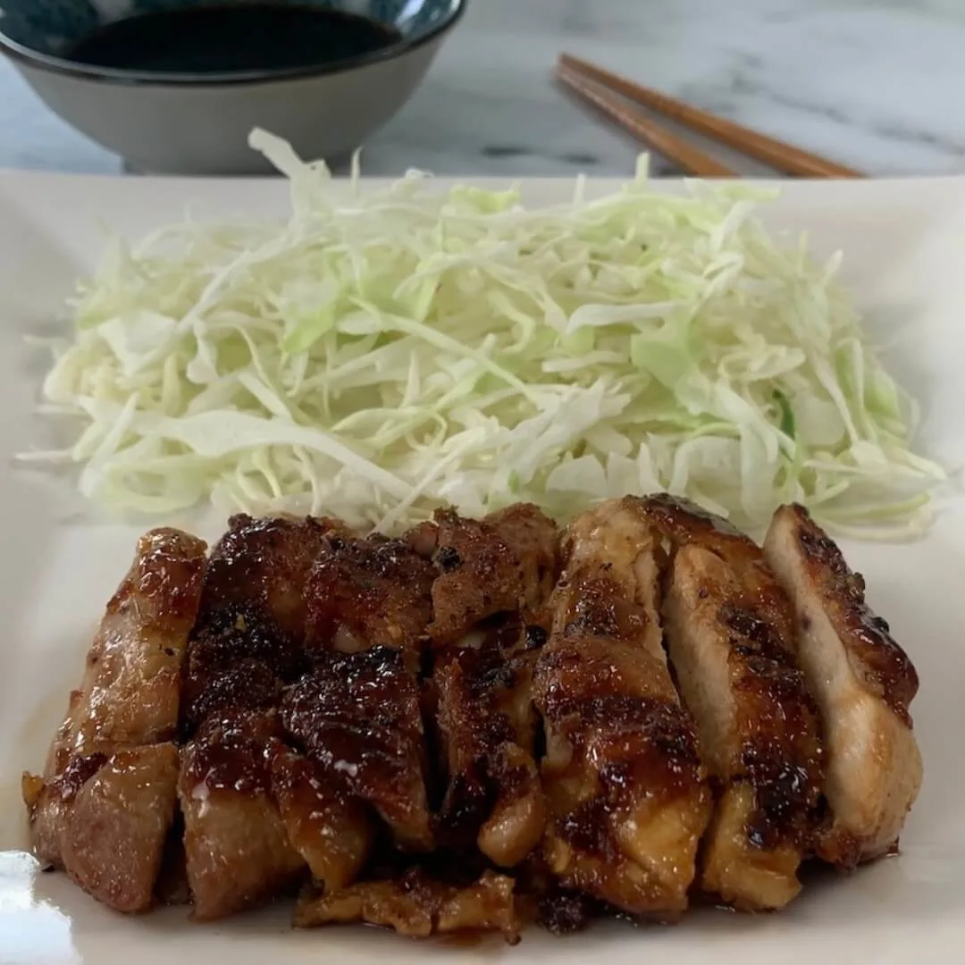 Chicken teriyaki square pic on a white plate with Japanese coleslaw and a ramekin of teriyaki sauce in the background.