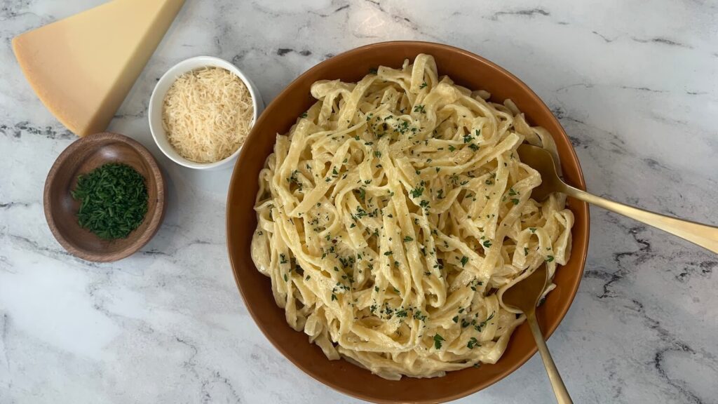 Alfredo sauce over fettuccini pasta in a brown bowl with ramekins of parmesan cheese and parsley, and block of cheese in the background. 