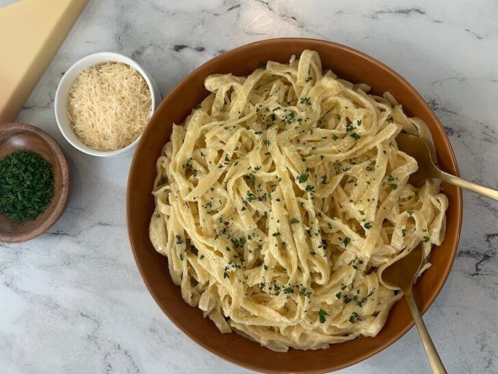 Alfredo sauce over fettuccini pasta in a brown bowl with ramekins of parmesan cheese and parsley, and block of cheese in the background.