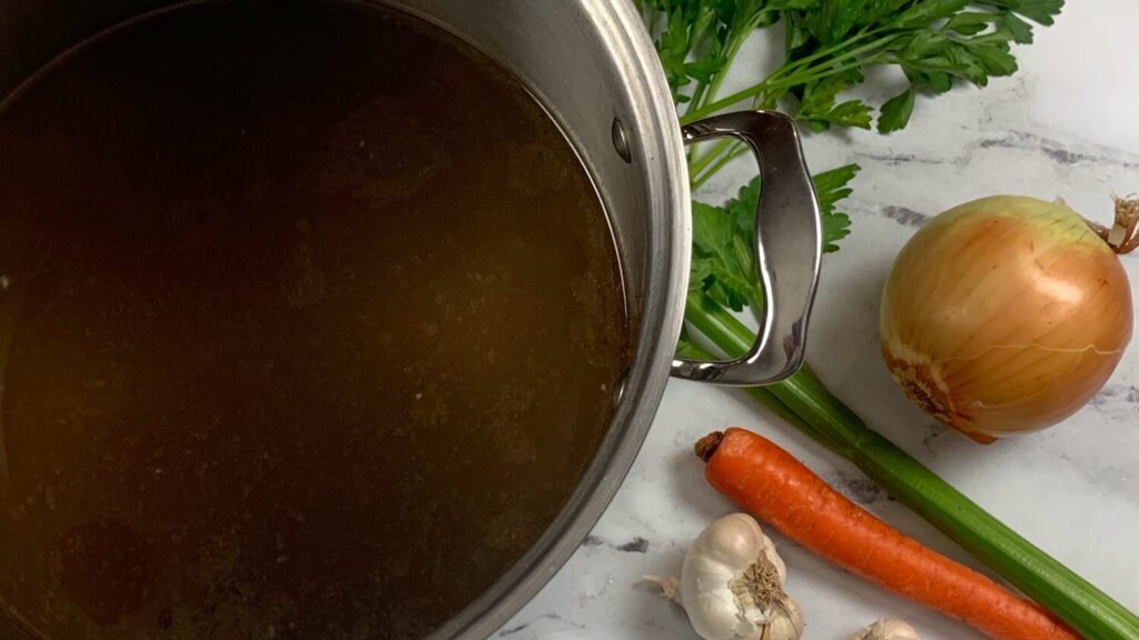 Beef broth in a pot with parsley, garlic, carrots, and celery in the background