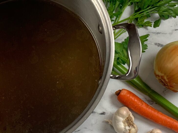 Beef broth in a pot with parsley, garlic, carrots, and celery in the background