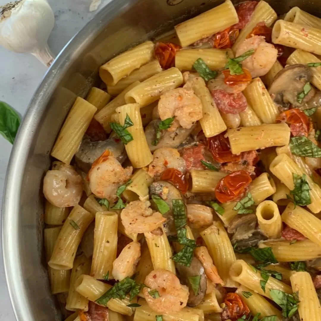 Boursin cheese pasta with shrimp in a large pan with bulbs of garlic and basil leaves in the background.