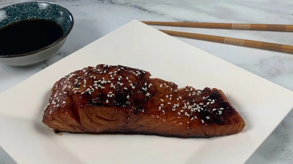 Teriyaki salmon horizontal pic on a white plate garnished with white sesame seeds.  Chopsticks and a ramekin of teriyaki sauce are in the background.