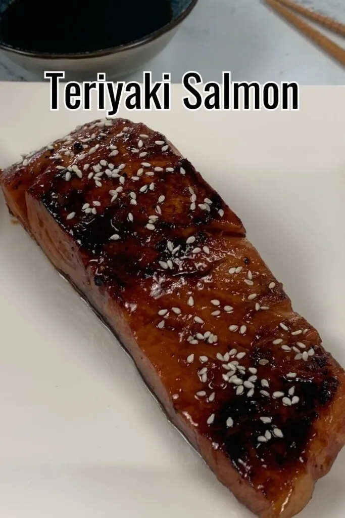 Teriyaki salmon garnished with white sesame seeds vertical shot.  Presented on a white plate with a ramekin of teriyaki sauce in the background.