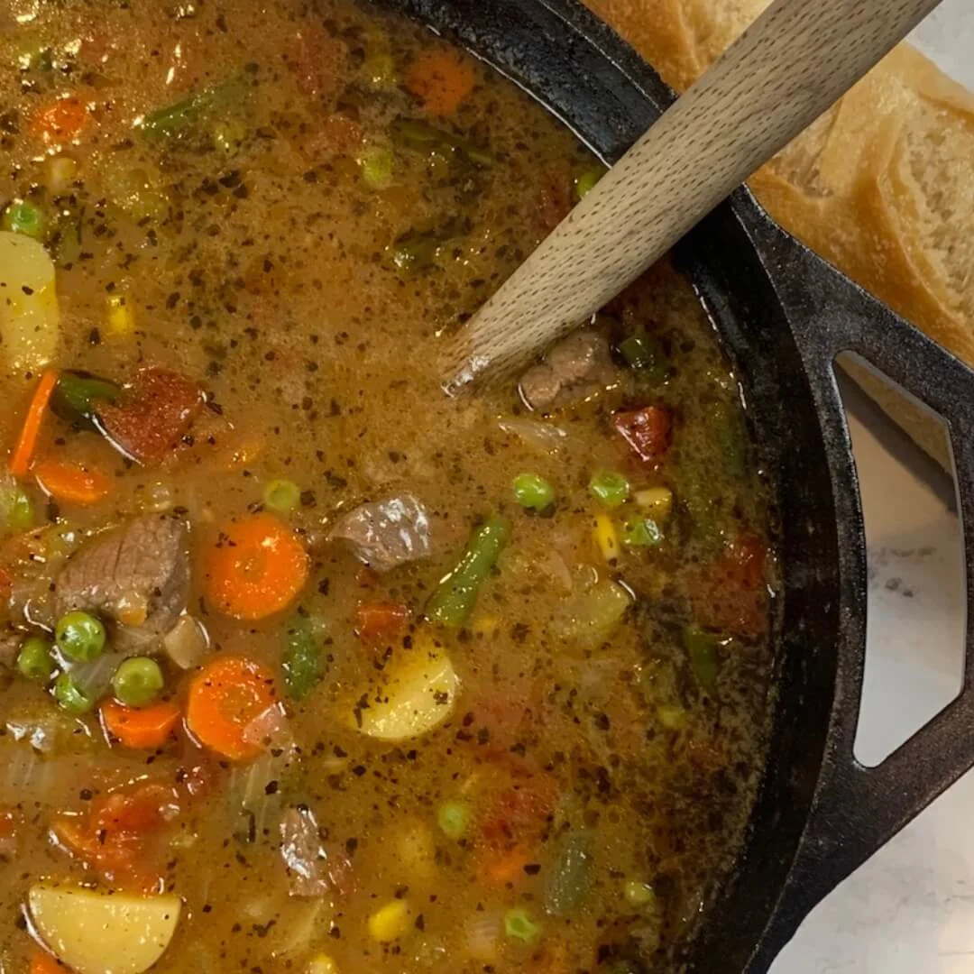 Homemade vegetable beef soup in a large cast iron pot square pic. Shown with a loaf of sourdough bread in the background.
