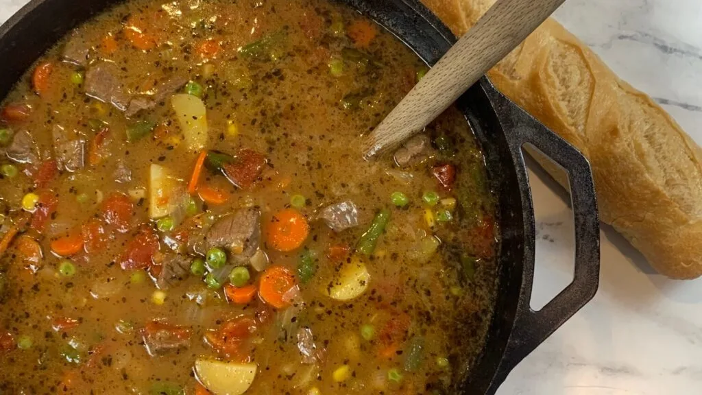 Horizontal vegetable beef soup pic. Shown in a cast iron pot and with a loaf of bread in the background.